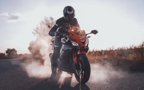 Motocyklista w kasku i kombinezonie na pomarańczowym motocyklu Barton Blade, z opon dymiących na drodze.
