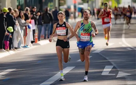 Atleti si preparano a una maratona, correndo fianco a fianco. La folla incita lungo il percorso.