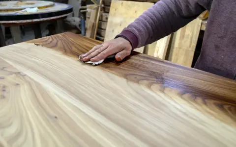 Mano che applica la finitura a cera dopo impregnante su un tavolo di legno grezzo, esaltandone le venature.