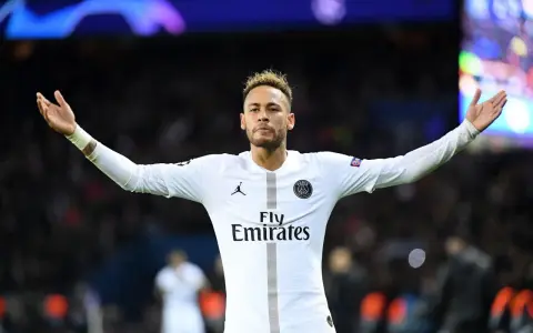 Neymar w białej koszulce PSG, z charakterystyczną neymar fryzurą, unosi ręce w geście radości na tle rozmazanych świateł stadionu.