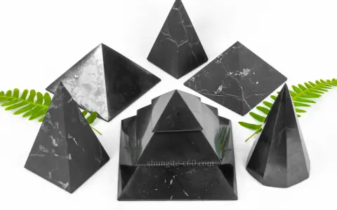 Seis pirámides de piedra shungita, algunas con vetas blancas, dispuestas sobre un fondo blanco con hojas verdes.