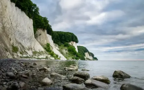 Majestatyczne, białe klify Rugii wznoszą się nad kamienistą plażą i spokojnym morzem. Zielone drzewa porastają szczyty, a chmury tworzą dramatyczne tło.