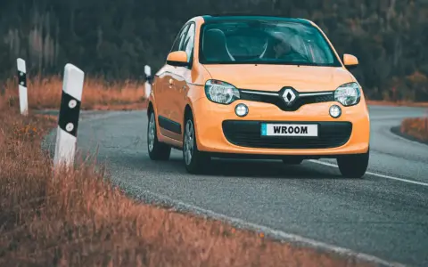 Une Twingo orange roule sur une route sinueuse bordée d'herbes sèches.