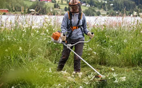 Persona con equipo de protección usa desbrozadora para cortar hierba alta, mostrando la diferencia entre desbrozadora y cortabordes en un paisaje rural.