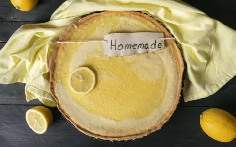Ein hausgemachter Zitronenkuchen mit einem Zitronenschnitz und einem "Homemade"-Etikett, umgeben von gelben Tüchern und Zitronen.
