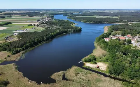 Malownicze jezioro Wojnowskie otoczone lasami i polami. W oddali widać zabudowania wsi, a na brzegu plażę z pomostem.