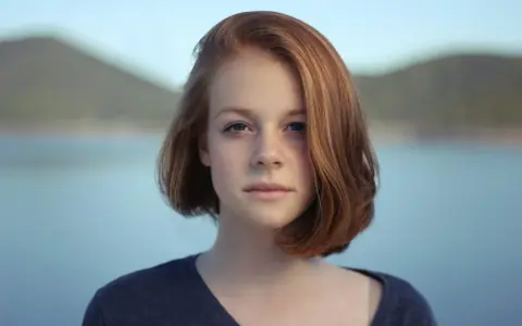 Ritratto fotografia di una giovane ragazza con capelli rossi e occhi azzurri, sullo sfondo di un lago e colline.