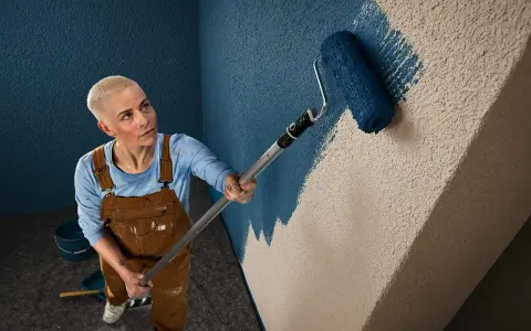 Frau mit Kurzhaarschnitt streicht mit blauem Roller eine Wand mit Rauputz.