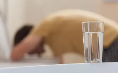 Person böjer sig över toaletten, troligen magsjuk av gammalt vatten. Ett glas vatten står på en vit yta.