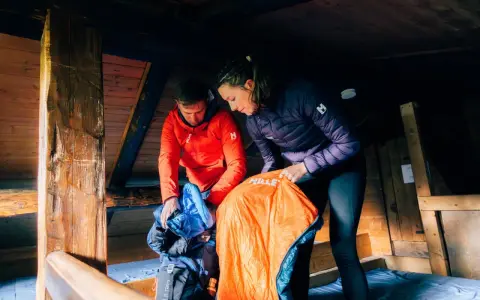 Préparation pour une nuit en refuge. Un homme en veste rouge et une femme en veste violette déballent des sacs de couchage dans une cabane en bois.