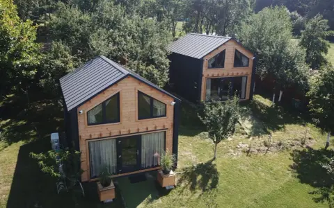 Dwa nowoczesne domki z drewna i czarnego panelu, idealne projekty domów pod wynajem, otoczone zielenią.