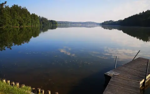 Spokojne jezioro Sulęczyno o poranku. Drewniane molo zaprasza do podziwiania odbijających się w tafli chmur i zielonych lasów.
