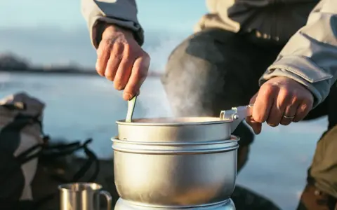 En person lagar mat utomhus, ångande kastruller och en mugg i metall antyder en lyxig matupplevelse i naturen.