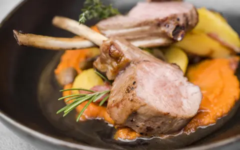 Chuleta de ternasco jugosa, servida sobre puré de zanahoria y patatas asadas, decorada con romero y tomillo.
