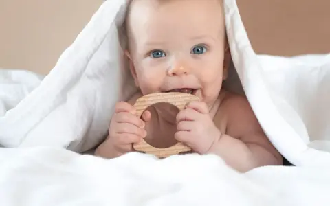 Ein Baby mit blauen Augen kaut auf einem Holzring. Vielleicht ist das der erster zahn baby, der durchbricht.