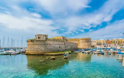 A stone castle overlooks a harbor filled with boats and yachts under a bright blue sky.