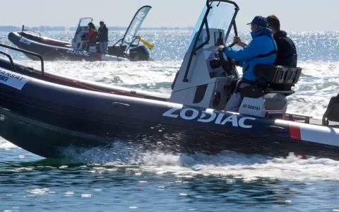 Deux bateaux Zodiac naviguent sur l'eau. L'un d'eux, au premier plan, porte le logo "ZODIAC" et semble communiquer sur le canal 9 VHF.