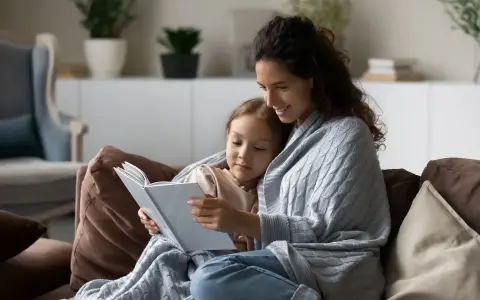 Mama czyta dziecku bajkę, otuleni kocem. Widać, ile jest opieki na dziecko w tej chwili.