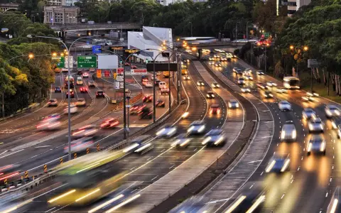 Traffico intenso su un'autostrada urbana al tramonto. Le luci dei veicoli creano scie luminose, con un **segnale 105** che indica la direzione.