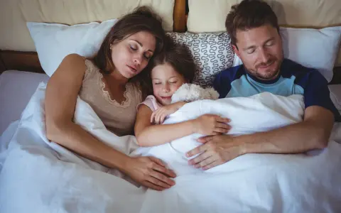 Familia durmiendo junta, la niña abraza un peluche. Un momento de paz que evoca la regresión infantil y la seguridad del hogar.
