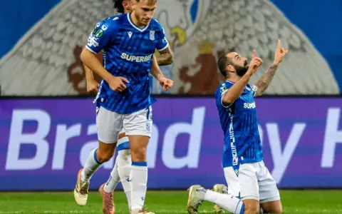 Piłkarz klęczy na boisku, celebrując bramkę. W tle biegnie kolega z drużyny. Wszyscy w niebieskich strojach. Czekamy, kiedy wraca ekstraklasa!