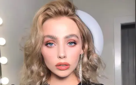 Kobieta z pięknymi, falowanymi włosami w odcieniu **naturalny średni blond**. Jej oczy podkreślone są delikatnym makijażem, a usta w kolorze brzoskwini.