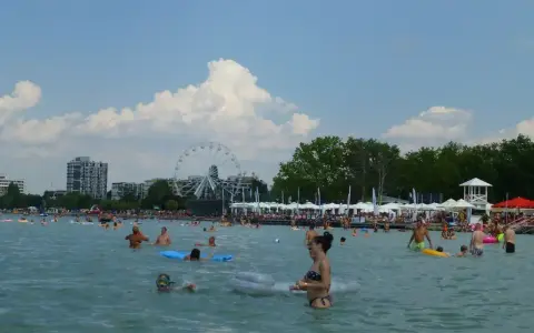Słoneczna balaton plaża tętni życiem. Ludzie kąpią się, pływają na materacach i odpoczywają pod parasolami. W tle widać diabelski młyn.