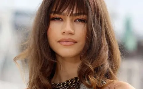Primo piano di una donna con un **paffuto taglio capelli viso tondo**. Capelli castani mossi con frangia, labbra lucide e una collana a catena spessa.