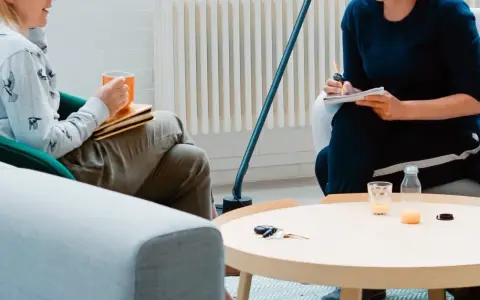 Dwie osoby siedzą w fotelach, jedna trzyma kubek, druga pisze w notatniku.