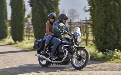 Coppia su una Moto Guzzi V7, con passeggero, percorre una strada alberata in una giornata di sole.