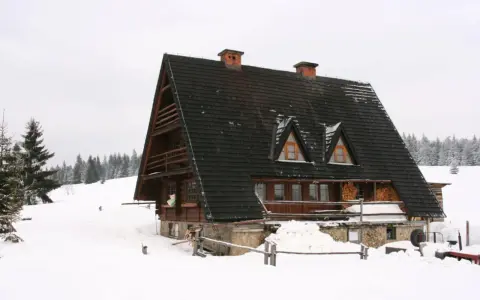 Drewniana bacówka w zimowej scenerii, otoczona śniegiem i lasem.