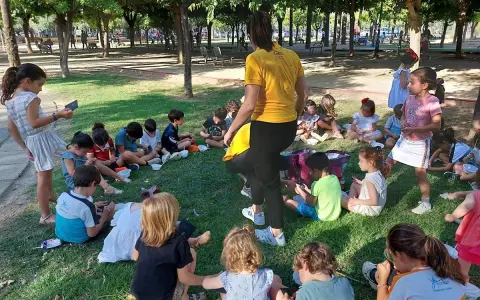 Niños sentados en el césped, participando en una gymkana para niños. Un adulto con camiseta amarilla dirige la actividad.