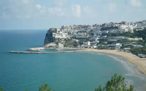 En vit stad klättrar uppför en klippa vid havet i Puglia, Italien. En strand och en hamn syns nedanför.