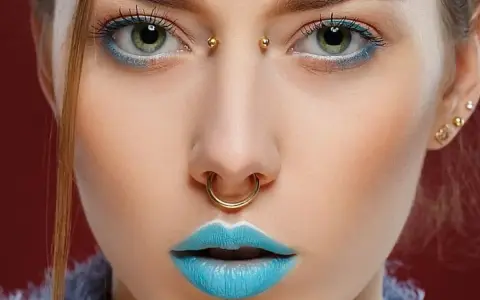 Kobieta z błękitnymi ustami i złotym septum. Jej oczy podkreślone są niebieskim cieniem, a między brwiami widoczny jest bridge piercing.