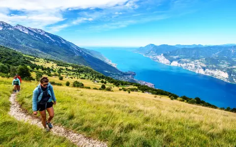 Zwei Wanderer genießen die Aussicht beim Monte Baldo Wandern. Der Weg führt über einen sonnigen Hang mit Blick auf den tiefblauen Gardasee.