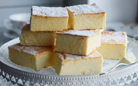 Sernik bez spodu, który zawsze się udaje, pokrojony w kostkę i posypany cukrem pudrem, ułożony na paterze.