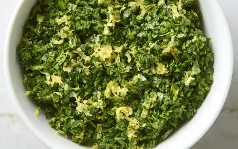 Un cuenco blanco lleno de perejil picado y ralladura de limón, perfecto para un toque fresco en tu cocina, como la albahaca en francés.