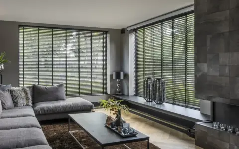 Nowoczesny salon z szarym narożnikiem i stolikiem kawowym. Duże okna zasłonięte czarnymi żaluzjami wewnętrznymi, które dodają elegancji.