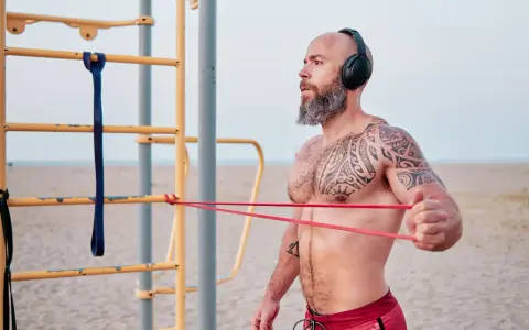Man med tatueringar och skägg tränar med gummisnoddar vid ett utegym på stranden.