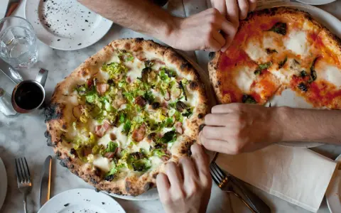 Dwie pizze na stole, jedna z brukselką i boczkiem, druga margherita. Ludzie sięgają po kawałki, pokazując, ile wyrasta ciasto na pizze.