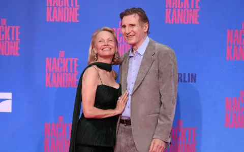 Liam Neeson e Pamela Anderson sorridono sul tappeto blu, con lo sfondo di "Die Nackte Kanone".