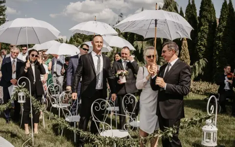Wesele w kwietniu pod białymi parasolami. Goście w eleganckich strojach, wśród nich pan młody z bukiecikiem.