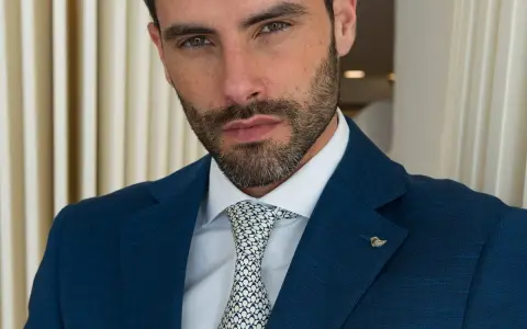 Uomo con abito blu, camicia bianca e cravatta con fantasia a pois bianchi e neri. Un tocco di stile con un pin a forma di ala.