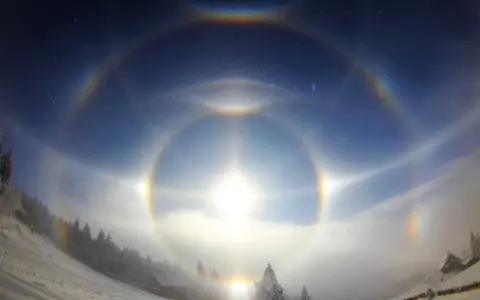 Zjawiskowy efekt halo nad zimowym krajobrazem. Słońce otoczone tęczowymi kręgami, mgła i ośnieżone drzewa tworzą magiczną scenę.