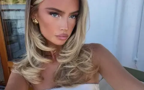 Kobieta z blond włosami, stylizowanymi na fale, z subtelnym makijażem i złotymi kolczykami. Jej fryzura przypomina buzz cut, ale jest dłuższa i bardziej objętościowa.
