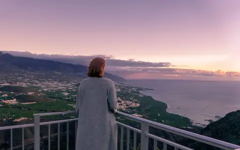 Frau blickt auf weite Landschaft, die ihr Angst vor Urlaub macht. Berge, Meer und Felder im Abendlicht.
