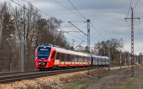 Czerwono-biały pociąg SKM S2 Chopin Airport jedzie po torach. System monitoringu ruchu pociągów śledzi jego trasę.