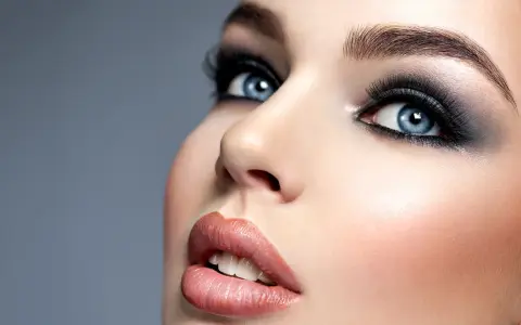Niebieskie oczy z perfekcyjnym makijażem smokey eye, podkreślone długimi rzęsami i błyszczącymi ustami.