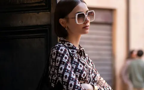 Emily in Paris, Lily Collins poses with large white sunglasses and a patterned shirt, leaning against a dark door.