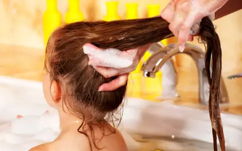 Schaumige Hände waschen lange, verfilzte Haare von Kindern in der Badewanne. Gelbe Flaschen im Hintergrund.